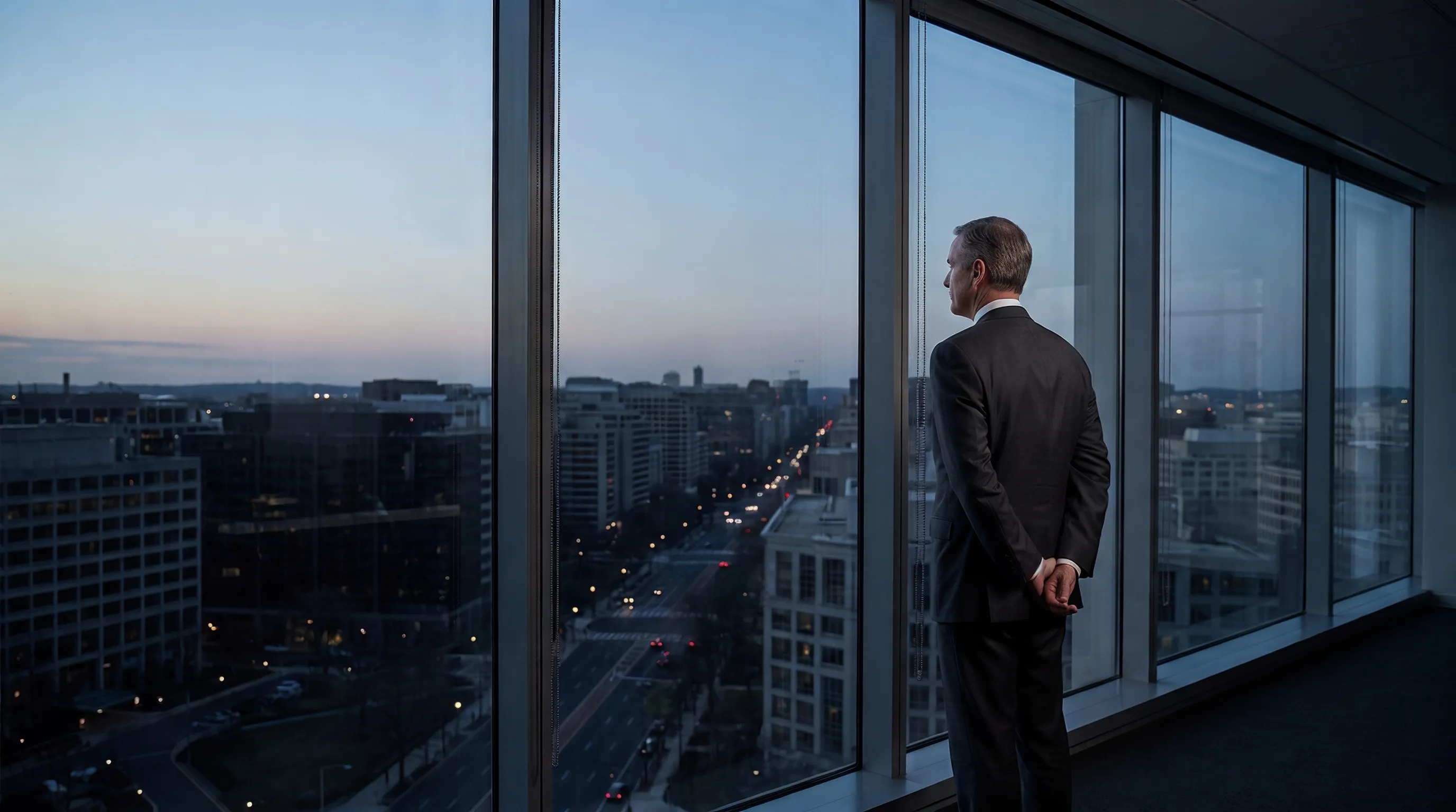 An executive in a dark suit stands with hands clasped behind his back, looking out a floor-to-ceiling office window at a city skyline as dusk settles and street lights come on below.